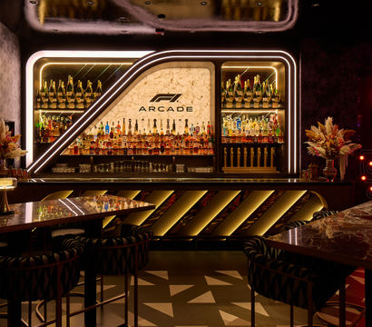 Neon-lit luxury bar interior with backlit marble bottle display, rows of spirits and champagne, marble tables, floral centerpieces and a glowing arcade gaming kiosk.