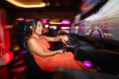 Smiling woman in an orange dress driving an arcade racing simulator with a steering wheel and large curved screen, colorful neon lights and motion blur in a lively gaming lounge.