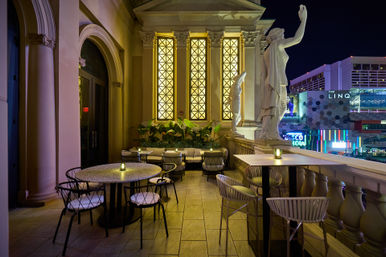 Candlelit rooftop terrace with classical marble statues, arched glowing windows and cozy seating overlooking a neon-lit Las Vegas skyline at night.