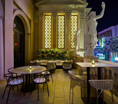 Candlelit rooftop terrace with classical marble statues, arched glowing windows and cozy seating overlooking a neon-lit Las Vegas skyline at night.
