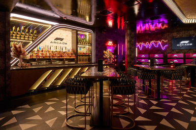 Neon-lit arcade bar interior with illuminated bottle wall, marble high-top tables, patterned stools and gaming screens in moody purple and gold lighting