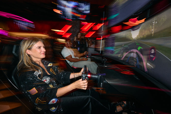 Woman in a racing cockpit playing an indoor arcade driving simulator, hands on a steering wheel with a large curved screen showing a virtual racetrack, neon red and purple lights and motion blur creating a fast-paced arcade atmosphere