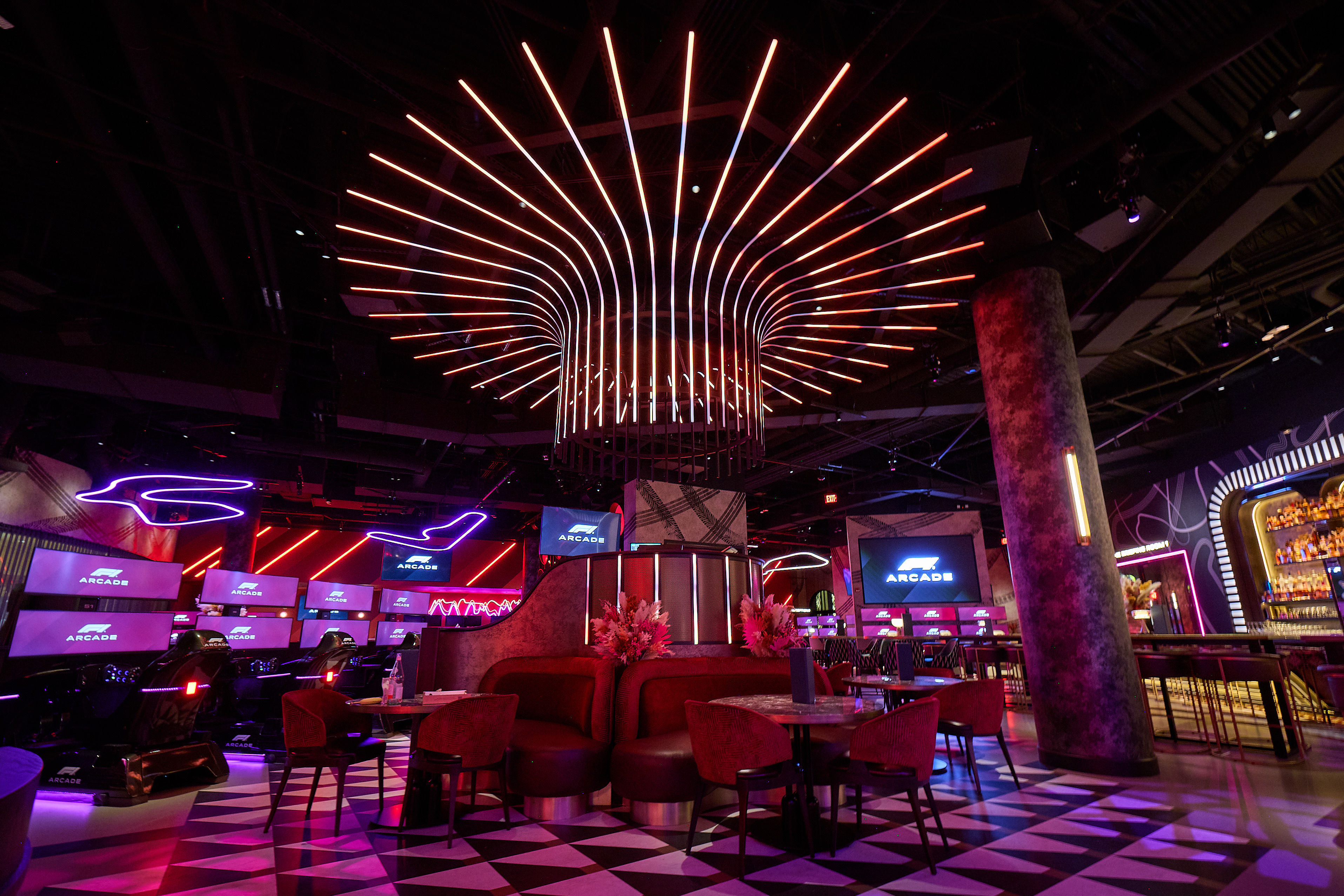 Neon-lit gaming lounge interior with a radial LED ceiling sculpture, red velvet booths, racing simulators and multiple arcade screens over a black-and-white geometric floor.