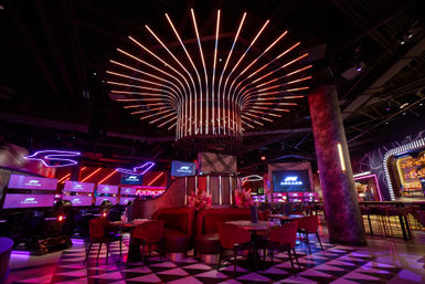 Neon-lit gaming lounge interior with a radial LED ceiling sculpture, red velvet booths, racing simulators and multiple arcade screens over a black-and-white geometric floor.