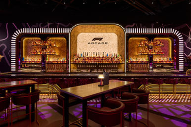 Neon-lit arcade-style cocktail bar interior with curved illuminated backbar, marble central panel, shelves packed with liquor bottles, high tables and plush stools.