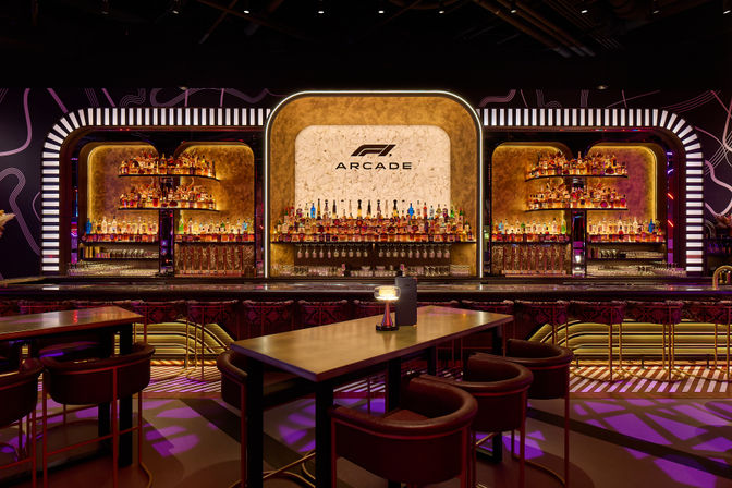 Neon-lit arcade-style cocktail bar interior with curved illuminated backbar, marble central panel, shelves packed with liquor bottles, high tables and plush stools.