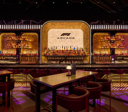 Neon-lit arcade-style cocktail bar interior with curved illuminated backbar, marble central panel, shelves packed with liquor bottles, high tables and plush stools.