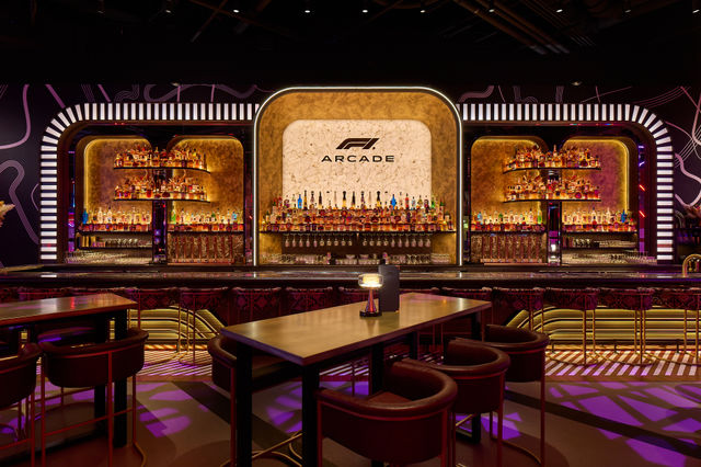 Neon-lit arcade-style cocktail bar interior with curved illuminated backbar, marble central panel, shelves packed with liquor bottles, high tables and plush stools.