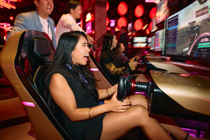 Woman laughing while driving a neon-lit arcade racing simulator with friends cheering and a large screen racing game in an indoor gaming arcade
