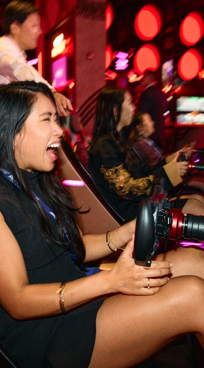 Woman laughing while driving a neon-lit arcade racing simulator with friends cheering and a large screen racing game in an indoor gaming arcade