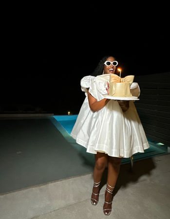 Celebrant in a short white puffy dress and round white sunglasses holding a single‑candle cake by a glowing pool at night
