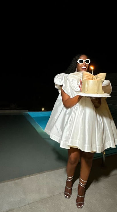 Celebrant in a short white puffy dress and round white sunglasses holding a single‑candle cake by a glowing pool at night