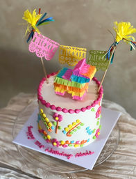 Fiesta-ready single-tier white birthday cake with bright piped flowers and pink bead border, topped by a tiny rainbow piñata, colorful papel picado banner and tissue tassels.