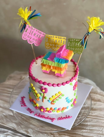 Fiesta-ready single-tier white birthday cake with bright piped flowers and pink bead border, topped by a tiny rainbow piñata, colorful papel picado banner and tissue tassels.