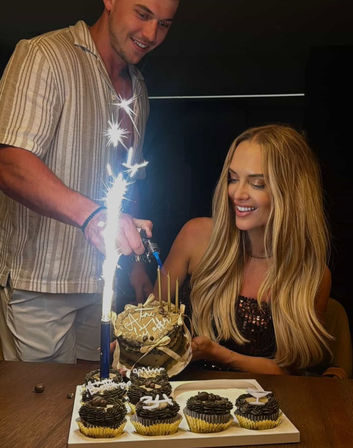 Indoor birthday celebration with a smiling woman holding a small layered cake while a man lights a tall sparkler candle; chocolate cupcakes in gold liners on a wooden table.