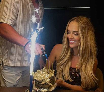 Indoor birthday celebration with a smiling woman holding a small layered cake while a man lights a tall sparkler candle; chocolate cupcakes in gold liners on a wooden table.
