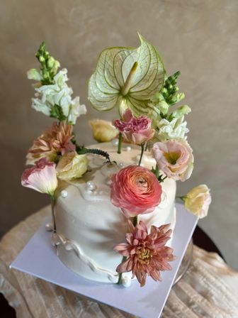 White fondant single-tier cake adorned with fresh blush ranunculus, peach dahlia, pale lisianthus, snapdragons and a green anthurium leaf, dotted with pearl accents — elegant floral celebration cake.