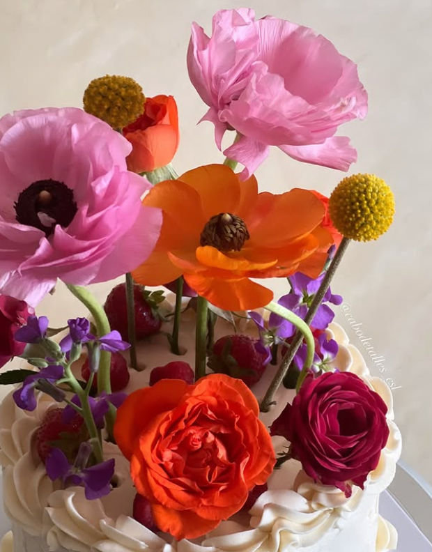 White buttercream cake with a playful floral topper of vibrant pink and orange blooms, yellow billy balls, purple accents and fresh strawberries