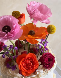 White buttercream cake with a playful floral topper of vibrant pink and orange blooms, yellow billy balls, purple accents and fresh strawberries