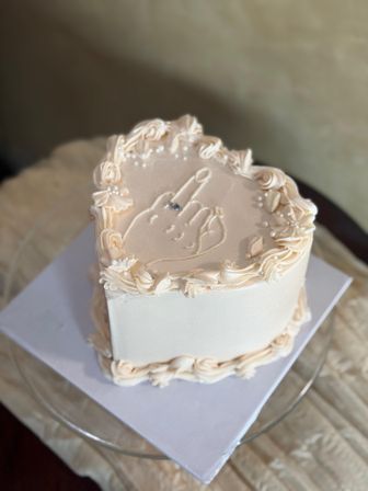 Close-up of a pale peach heart-shaped buttercream novelty cake on a glass stand, decorated with rosette borders, edible pearls and a piped hand giving the middle finger.