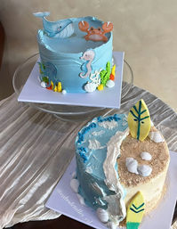 Two ocean-themed celebration cakes: a pastel blue cake topped with a whale, crab, seahorse and octopus with seaweed and shell accents, and a half-beach, half-ocean cake with sandy crumbs, surfboard toppers, waves and edible shells.
