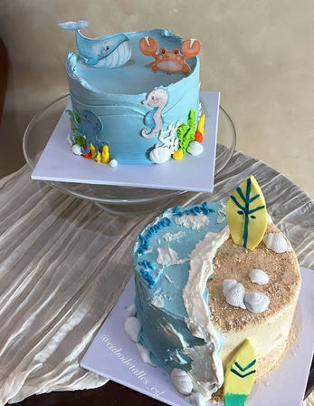Two ocean-themed celebration cakes: a pastel blue cake topped with a whale, crab, seahorse and octopus with seaweed and shell accents, and a half-beach, half-ocean cake with sandy crumbs, surfboard toppers, waves and edible shells.