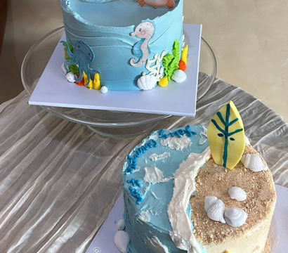 Two ocean-themed celebration cakes: a pastel blue cake topped with a whale, crab, seahorse and octopus with seaweed and shell accents, and a half-beach, half-ocean cake with sandy crumbs, surfboard toppers, waves and edible shells.