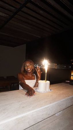 Smiling woman on a rooftop terrace at night with a white frosted cake topped by a tall sparkler and city lights in the background.