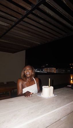 Person in a white one-shoulder dress laughing at a rooftop terrace at night with a small white cake topped by a tall gold candle and city skyline lights in the background.