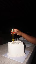 Hand with bright red nails placing a gold sparkler candle into a small white frosted celebration cake on a marble countertop against a dark background