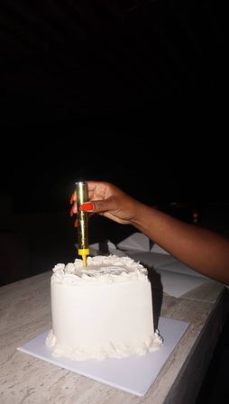 Hand with bright red nails placing a gold sparkler candle into a small white frosted celebration cake on a marble countertop against a dark background