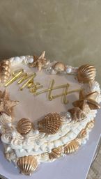 Beach-inspired ivory buttercream cake decorated with gold edible seashells and starfish, pearl accents, piped rosettes and gold script