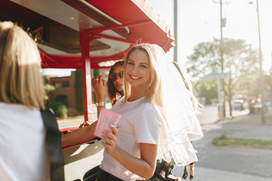 Sunlit bachelorette scene: smiling bride-to-be in a veil and white tee holding a pink cup aboard a red pedal party bike on a city street.