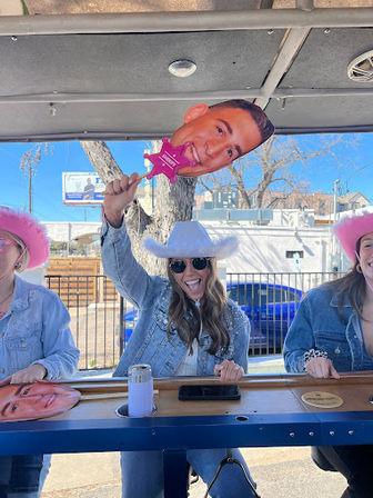 Three friends in denim jackets and cowgirl hats laughing at an outdoor bar on a sunny day; center woman in a white hat and sunglasses holds a pink sheriff-star prop with a large face cutout above her head.