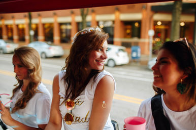Three smiling women on a downtown city street outing; center woman wears a floral headband, white tee with gold text, sunglasses and a temporary ring tattoo on her upper arm, brick buildings and parked cars blurred in the background.