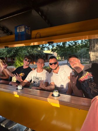 Four friends smiling and flashing hand signs while seated at a bright yellow pedal bar with canned drinks, trees and a wooden fence in a sunlit outdoor setting.