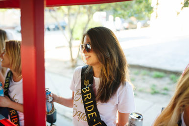 Smiling bride-to-be in sunglasses wearing a black 'BRIDE' sash, holding a canned drink on a sunny bachelorette party ride along a city sidewalk.