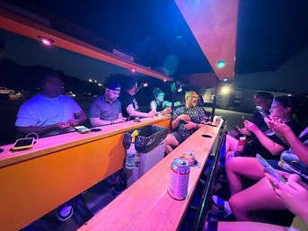Nighttime outdoor mobile bar scene with people seated along a wooden counter under colorful LED lights, canned drinks and phones on the bar, passengers chatting and using smartphones for a lively nightlife ride.