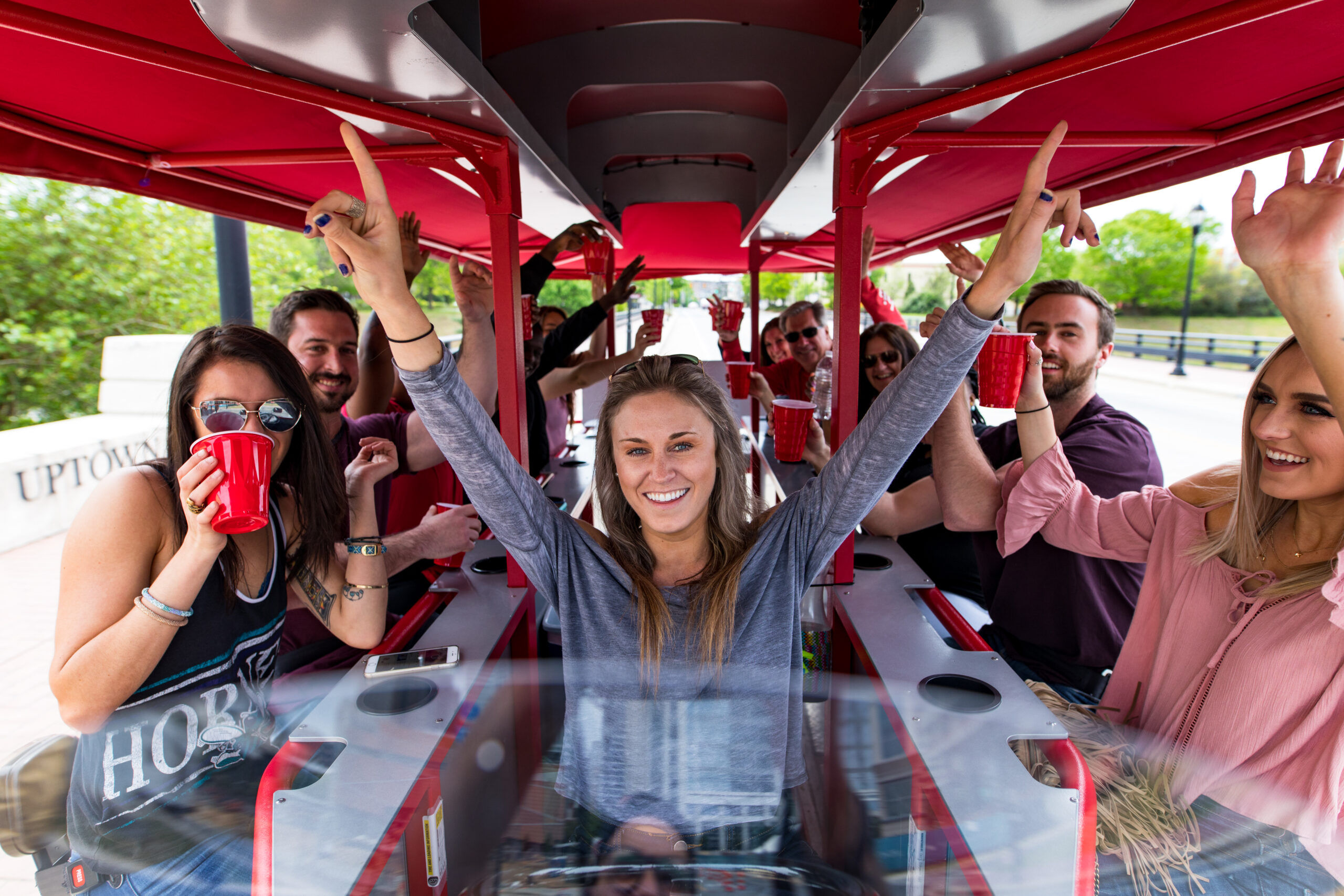 Smiling group of friends on a red‑canopied pedal pub party bike, cheering with red cups as a woman at the center raises her arms on an outdoor riverside/urban tour.