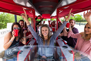 Smiling group of friends on a red‑canopied pedal pub party bike, cheering with red cups as a woman at the center raises her arms on an outdoor riverside/urban tour.