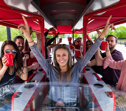 Smiling group of friends on a red‑canopied pedal pub party bike, cheering with red cups as a woman at the center raises her arms on an outdoor riverside/urban tour.