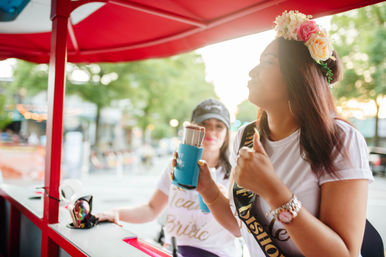 Two friends riding a pedal-powered party cart on a downtown street at sunset, one wearing a floral crown and bachelorette sash while holding a canned drink in a blue koozie and giving a thumbs-up.