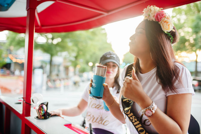 Two friends riding a pedal-powered party cart on a downtown street at sunset, one wearing a floral crown and bachelorette sash while holding a canned drink in a blue koozie and giving a thumbs-up.
