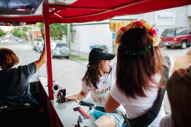 Friends cheering on a red pedal-powered party cart along a tree-lined city street; a woman in a white tee and baseball cap laughs while another wears a floral crown.