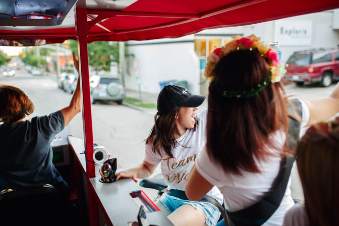 Friends cheering on a red pedal-powered party cart along a tree-lined city street; a woman in a white tee and baseball cap laughs while another wears a floral crown.