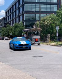 Bright blue sports car with white racing stripes on a sunny downtown street, passing a glass-front office building, trees, and a pedal-powered party bike with riders.