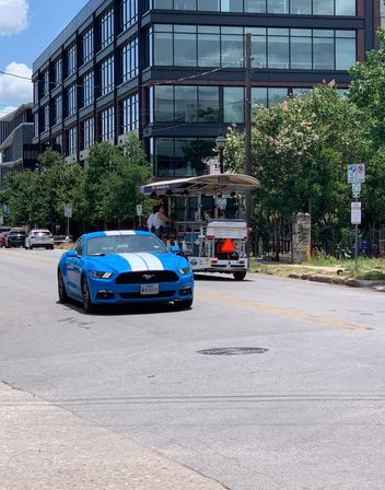 Bright blue sports car with white racing stripes on a sunny downtown street, passing a glass-front office building, trees, and a pedal-powered party bike with riders.