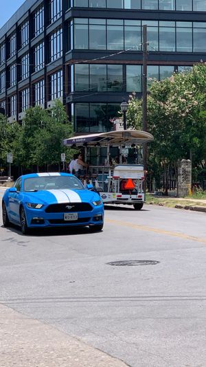 Bright blue sports car with white racing stripes on a sunny downtown street, passing a glass-front office building, trees, and a pedal-powered party bike with riders.