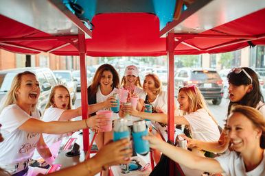 Smiling women on a red-canopied party bike in a downtown street, wearing matching shirts and cheering with cans and pink cups for a bachelorette-style toast.