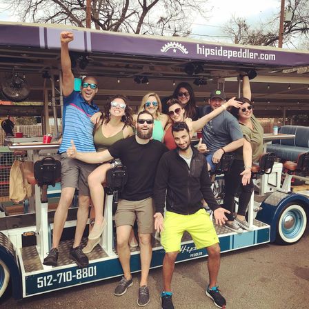 Energetic group of friends on a colorful mobile party bike/pedal bar, cheering and posing on bar stools and platform in casual summer clothes and sunglasses, parked on a tree-lined street